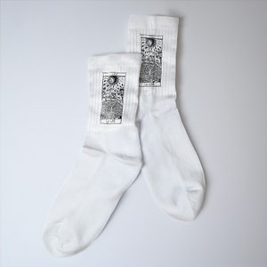 Può includere: Un paio di calzini bianchi con un design di carta dei tarocchi in bianco e nero. La carta è la carta della Luna, e il testo sulla carta recita "La Lune".