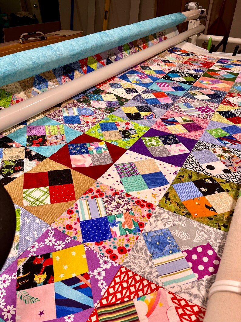 Pode incluir: Um cobertor com um design de patchwork que apresenta uma variedade de tecidos coloridos. O cobertor est&aacute; sendo acolchoado em uma m&aacute;quina de acolchoar de bra&ccedil;o longo.