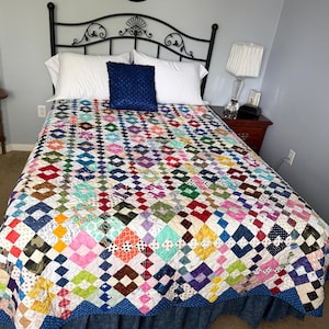 May include: A patchwork quilt featuring a repeating square pattern in a wide variety of colours and fabric prints, including red, blue, yellow, green, and pink. The quilt is draped over a bed with two white pillows and one dark blue pillow.