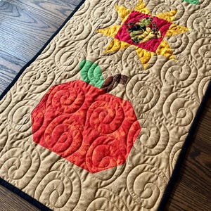 Peut inclure: Chemin de table matelassé avec une grande pomme rouge et un motif d'éclat d'étoile jaune. Le chemin de table a un fond beige avec un motif en spirale et une bordure noire. La pomme et l'éclat d'étoile sont appliqués sur le tissu.