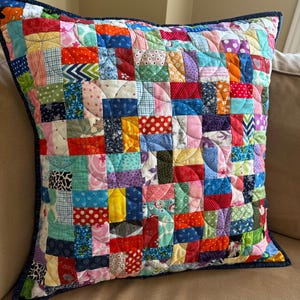 Peut inclure: Un coussin carré au motif patchwork composé de nombreuses petites pièces de tissu rectangulaires multicolores aux motifs variés, incluant des pois, des fleurs et des formes géométriques, bordé d'une bordure bleu foncé façon denim.