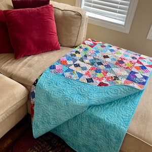Pode incluir: Um edredom azul turquesa com um padr&atilde;o floral est&aacute; dobrado em um sof&aacute; bege com almofadas vermelhas e bege. O edredom &eacute; feito de quadrados de patchwork em v&aacute;rias cores e padr&otilde;es.