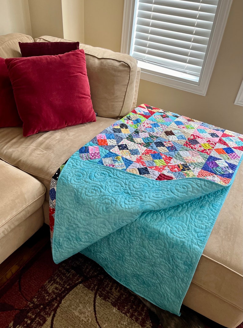 Pode incluir: Um edredom azul turquesa com um padr&atilde;o floral est&aacute; dobrado em um sof&aacute; bege com almofadas vermelhas e bege. O edredom &eacute; feito de quadrados de patchwork em v&aacute;rias cores e padr&otilde;es.