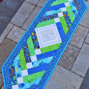 May include: A blue and green table runner with a white centre panel featuring a swirl design. The runner is decorated with a pattern of fish and bubbles.