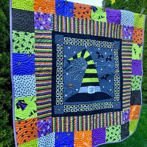 May include: A Halloween-themed quilt with a central design of a witch's hat, bats, and a spiderweb. The quilt features a patchwork design with squares of orange, green, purple, and black fabrics. The quilt is approximately 102 cm square.