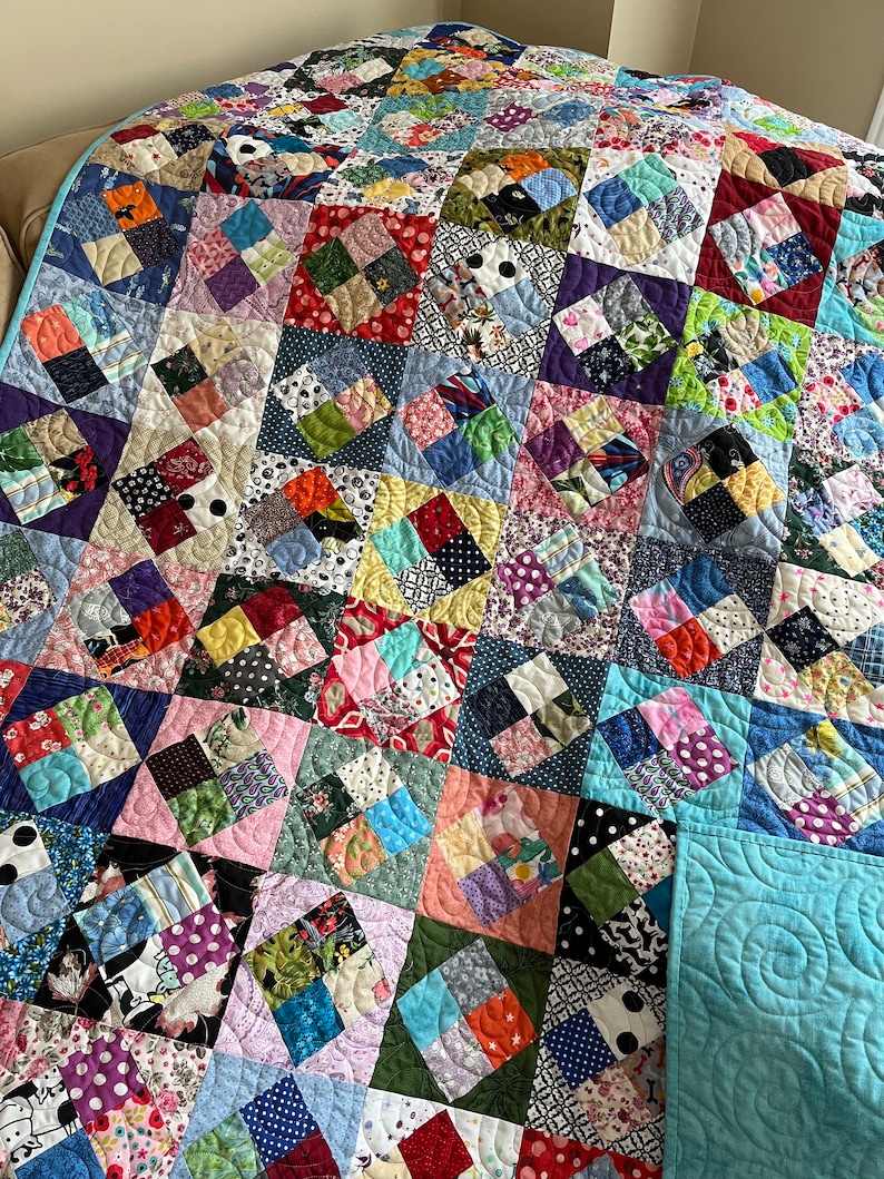 Pode incluir: Uma colcha de retalhos colorida com uma variedade de tecidos e padr&otilde;es. A colcha &eacute; feita de quadrados e diamantes, e &eacute; acolchoada com um padr&atilde;o de redemoinho. A colcha &eacute; de cor azul claro e est&aacute; dobrada ao meio.