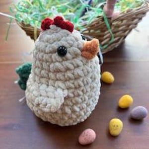Può includere: Un pollo all'uncinetto beige fatto a mano, con cresta rossa e becco arancione, è seduto accanto a un cestino di vimini pieno di erba di carta verde e bianca. Intorno al pollo sono sparsi quattro ovetti di Pasqua maculati gialli, rosa e viola.