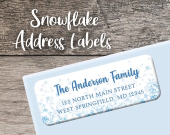 Op de afbeelding: Een blauw en wit sneeuwvlok-thema adreslabel met de tekst "The Anderson Family" en hun adres. Het label is op een lichtblauwe envelop met de tekst "Gedrukt of digitaal" op een blauw lint.