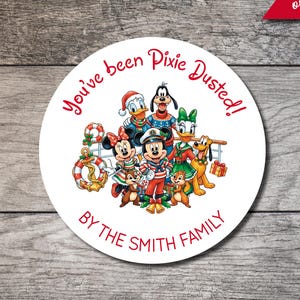 May include: A circular sticker featuring a group of cartoon characters in holiday attire, including Mickey, Minnie, Donald, Daisy, Goofy, Pluto, and Chip and Dale. The text reads You've been Pixie Dusted! and BY THE SMITH FAMILY in red.