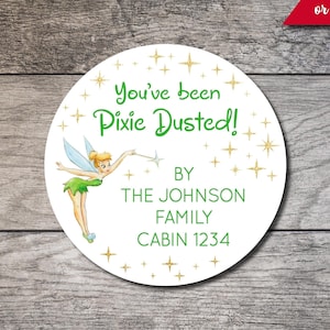 May include: A white round sticker with a green Tinkerbell illustration and gold glitter stars. The text reads "You've been Pixie Dusted!" with the words "BY THE JOHNSON FAMILY CABIN 1234" below.