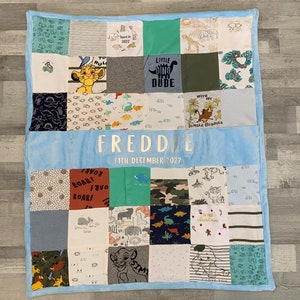 May include: A patchwork baby quilt with a light blue border and backing. The quilt is made from various fabric squares, some featuring animal and dinosaur graphics, and the name "FREDDIE" and date "11TH DECEMBER 2022" are embroidered on it.