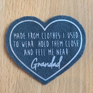May include: A grey felt heart-shaped keepsake with white text that reads "MADE FROM CLOTHES I USED TO WEAR HOLD THEM CLOSE AND FEEL ME NEAR Grandad".