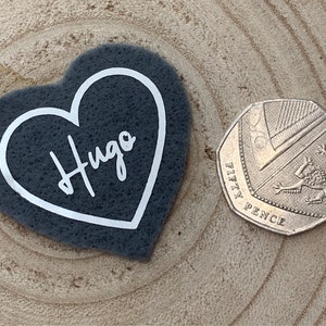 May include: A grey felt heart with a white outline and the name "Hugo" written in white inside. The heart is about the size of a 50 pence coin.