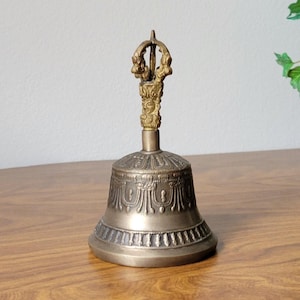 May include: A bronze-colored bell with intricate embossed patterns and a decorative handle. The bell has a flared base and a detailed handle with ornate carvings. The bell is likely used for religious or ceremonial purposes.