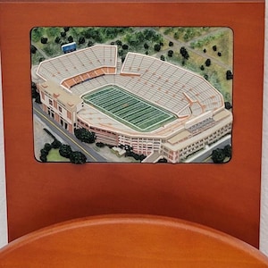 May include: A detailed, three-dimensional stadium model with a green field and tan seating. The model is set within a rectangular frame, mounted on a wooden surface. The stadium is surrounded by trees and roads.