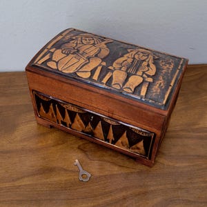 May include: A wooden box with a hinged lid and a small key. The lid features a copper-colored relief of two figures playing instruments. The front of the box has a copper-colored geometric design. The box is brown.