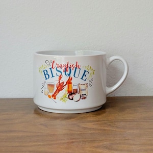 May include: White ceramic mug with a handle, featuring the words "Crawfish Bisque" in a colorful, decorative font. The design includes illustrations of a crawfish, ingredients, and a glass of beer. The mug sits on a wooden surface.