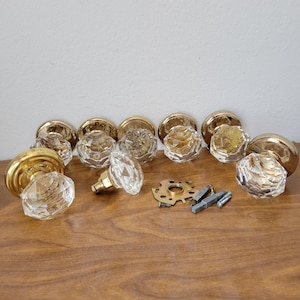 May include: A collection of vintage door knobs with clear, faceted glass and gold-toned metal bases. The hardware includes several complete knobs, along with mounting hardware and a decorative plate, all arranged on a wooden surface.