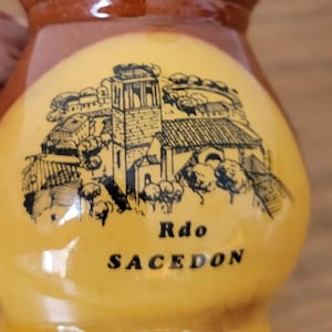 May include: A ceramic pitcher with a brown top and a yellow body. The yellow body features a black and white illustration of a church and the text "Rdo SACEDON".