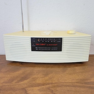 May include: A cream-colored, curved tabletop radio with a telescoping metal antenna. The front features a black display panel labeled THE CURVE by SUNTONE, showing FM and AM frequency scales, a power indicator light, and control sliders.