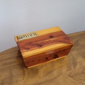 May include: A rectangular wooden box with a hinged lid. The box is made of cedar wood, with a lighter-colored top and darker sides. A gold-colored metal plate on the lid reads "MOTHER".