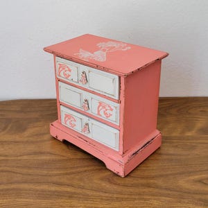 May include: A small coral-colored wooden chest with three white drawers. The top surface features a white mermaid graphic. Each drawer front is decorated with a coral-colored dolphin silhouette and a small, light-colored decorative knob.