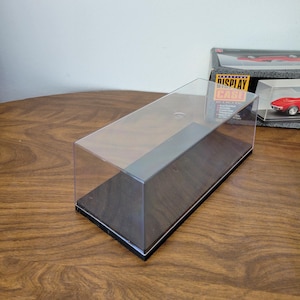 May include: A clear acrylic display case with a dark wood-grain base and a gray trim. The case is designed to showcase collectibles. The words "DISPLAY CASE" are visible on a box in the background.