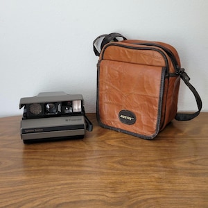 May include: A gray Polaroid Spectra System instant camera with a brown leather camera bag that says "RUSTIC" on it.