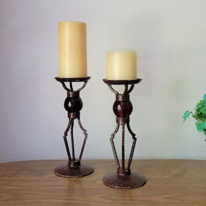 May include: Two decorative candle holders with cream-colored candles. The holders are made of dark brown metal with a decorative design, featuring a round base, a central glass orb, and a top platform for the candles. The candle holders are on a wooden surface.