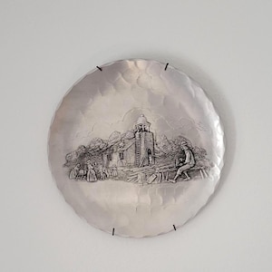May include: A silver metal wall plate with a raised design of a church building and people in a rural setting. The plate has a hammered texture and is hanging on a white wall.