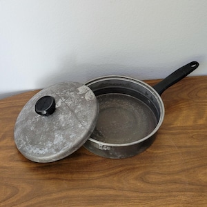 May include: A vintage aluminum saucepan with a black handle and a matching lid. The pan has a worn, gray finish.