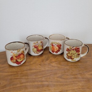 May include: Four ceramic mugs with fruit designs. Each mug features a different fruit illustration, including apples, pears, and grapes, in shades of red, yellow, and green. The mugs have a speckled beige finish with brown rims and handles.