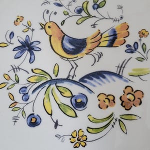 May include: A white ceramic plate with a hand-painted design featuring a yellow and blue bird perched on a blue hill. The bird is surrounded by blue, yellow, and orange flowers and green leaves. The design has a whimsical, folk art style.