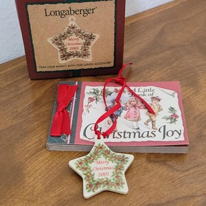May include: A star-shaped Christmas ornament with "Merry Christmas 2001" text and a holly wreath design. The ornament hangs from a red ribbon, next to a small book titled "A Little Book of Christmas Joy" and a Longaberger box.