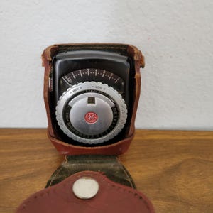 Puede incluir: Fotómetro GE vintage en una funda de cuero marrón. El fotómetro tiene una esfera negra con marcas blancas y plateadas, incluyendo un dial con ajustes de apertura y tiempo. La funda está abierta, revelando la esfera del fotómetro.
