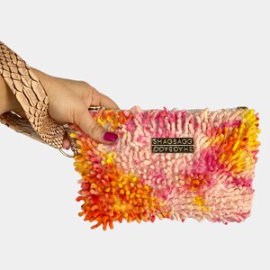 May include: A pink, orange, and yellow shaggy clutch with a gold metal tag that reads "SHAGBAGG".