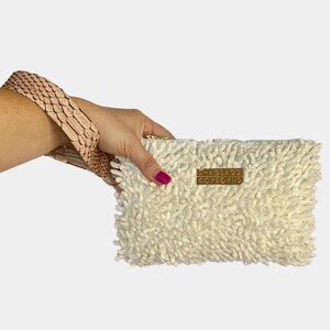 Può includere: Una pochette bianca e soffice con una cerniera dorata e un'etichetta in metallo dorato che dice "SHAGBAGG 9C7859HS".