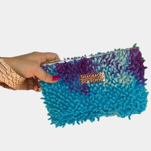 May include: A blue, purple and green shaggy fabric clutch with a gold zipper and the text "SHAGBAG" printed on the front.