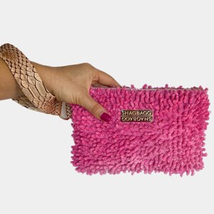 May include: A pink fluffy clutch with a gold metal logo that reads "SHAGBAGG" in all caps. The clutch is held in a hand with a brown and beige snake skin bracelet.