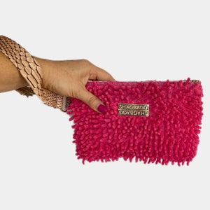 May include: A pink fluffy clutch with a gold metal tag that says "SHAGBAG" on it. The clutch has a brown leather strap.