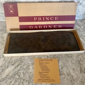 May include: A brown leather wallet in a white box with a brown and red box lid. The lid has the text "PRINCE GARDNER" in gold lettering. The box also includes a small card with the text "Princess Gardner" and "Prince Gardner" listing various leather goods.