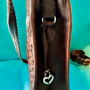 Puede incluir: Un bolso de mano de cuero marr&oacute;n con una correa trenzada y un dije en forma de coraz&oacute;n. El bolso tiene un dise&ntilde;o tejido y un cierre de solapa.