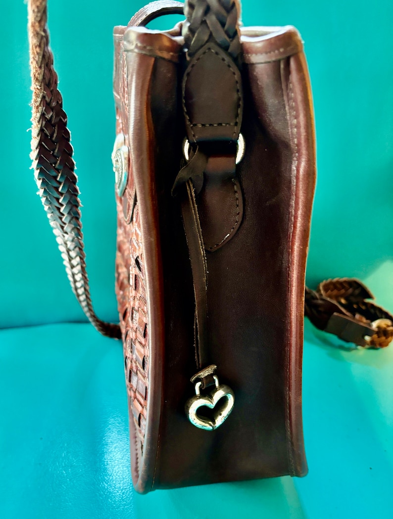 Puede incluir: Un bolso de mano de cuero marr&oacute;n con una correa trenzada y un dije en forma de coraz&oacute;n. El bolso tiene un dise&ntilde;o tejido y un cierre de solapa.
