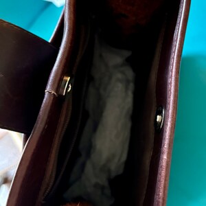 Puede incluir: El interior de un bolso de mano de cuero marr&oacute;n con cierre de cremallera y un bot&oacute;n plateado. El bolso est&aacute; vac&iacute;o y tiene un forro suave y texturizado.