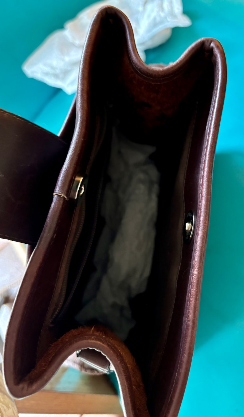 Puede incluir: El interior de un bolso de mano de cuero marr&oacute;n con cierre de cremallera y un bot&oacute;n plateado. El bolso est&aacute; vac&iacute;o y tiene un forro suave y texturizado.