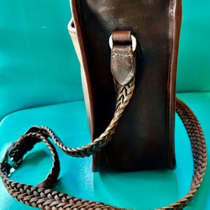 Puede incluir: Un bolso de cuero marr&oacute;n con correa trenzada. El bolso tiene forma rectangular y una hebilla plateada.