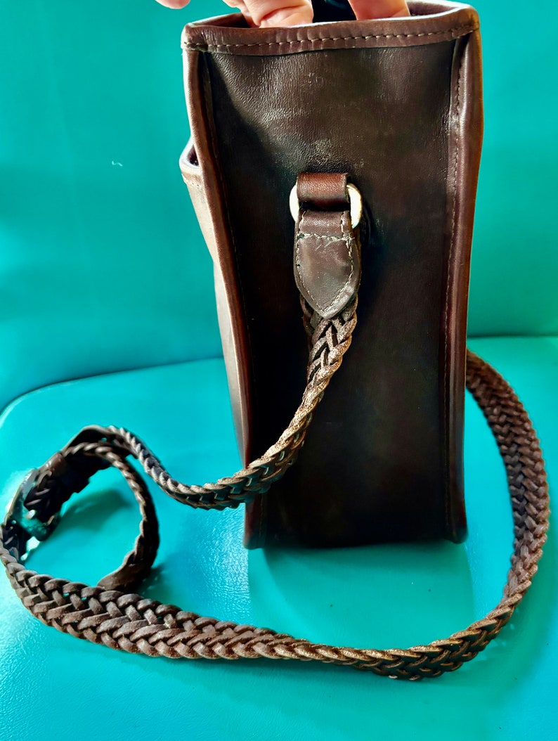 Puede incluir: Un bolso de cuero marr&oacute;n con correa trenzada. El bolso tiene forma rectangular y una hebilla plateada.