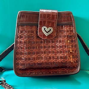 Puede incluir: Un bolso de mano de cuero marr&oacute;n con un patr&oacute;n tejido y un dije de coraz&oacute;n plateado. El bolso tiene una correa larga y un cierre de solapa.