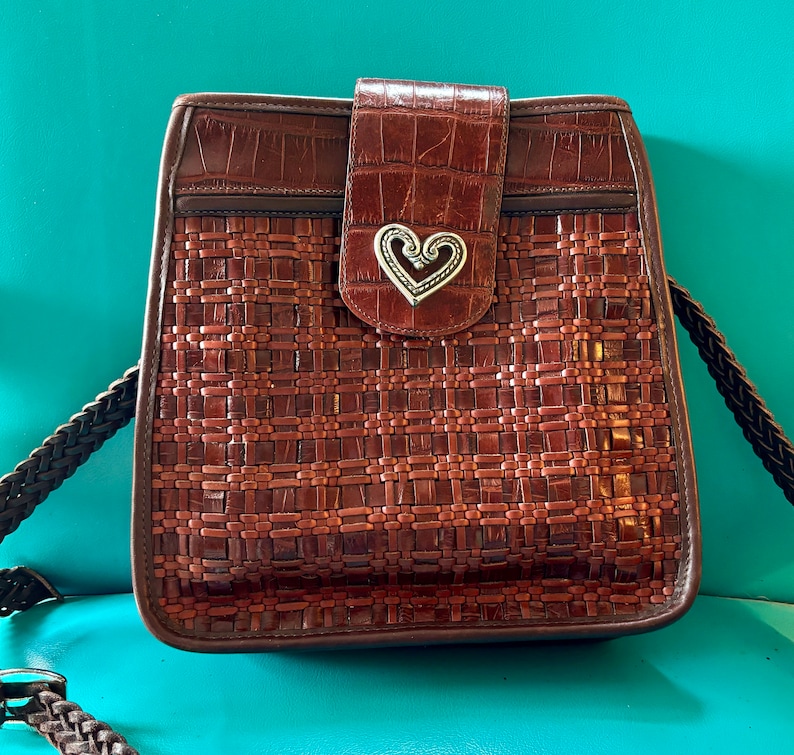 Puede incluir: Un bolso de mano de cuero marr&oacute;n con un patr&oacute;n tejido y un dije de coraz&oacute;n plateado. El bolso tiene una correa larga y un cierre de solapa.