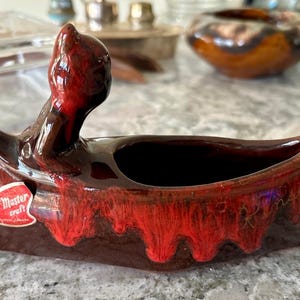 Könnte beinhalten: Eine rote und braune Keramik-Schale in Form eines Kanus mit roter Glasur und einem "Mr. Master Craft"-Etikett. Die Schale ist mit einer roten und braunen Glasur verziert und hat eine kleine Öffnung oben.